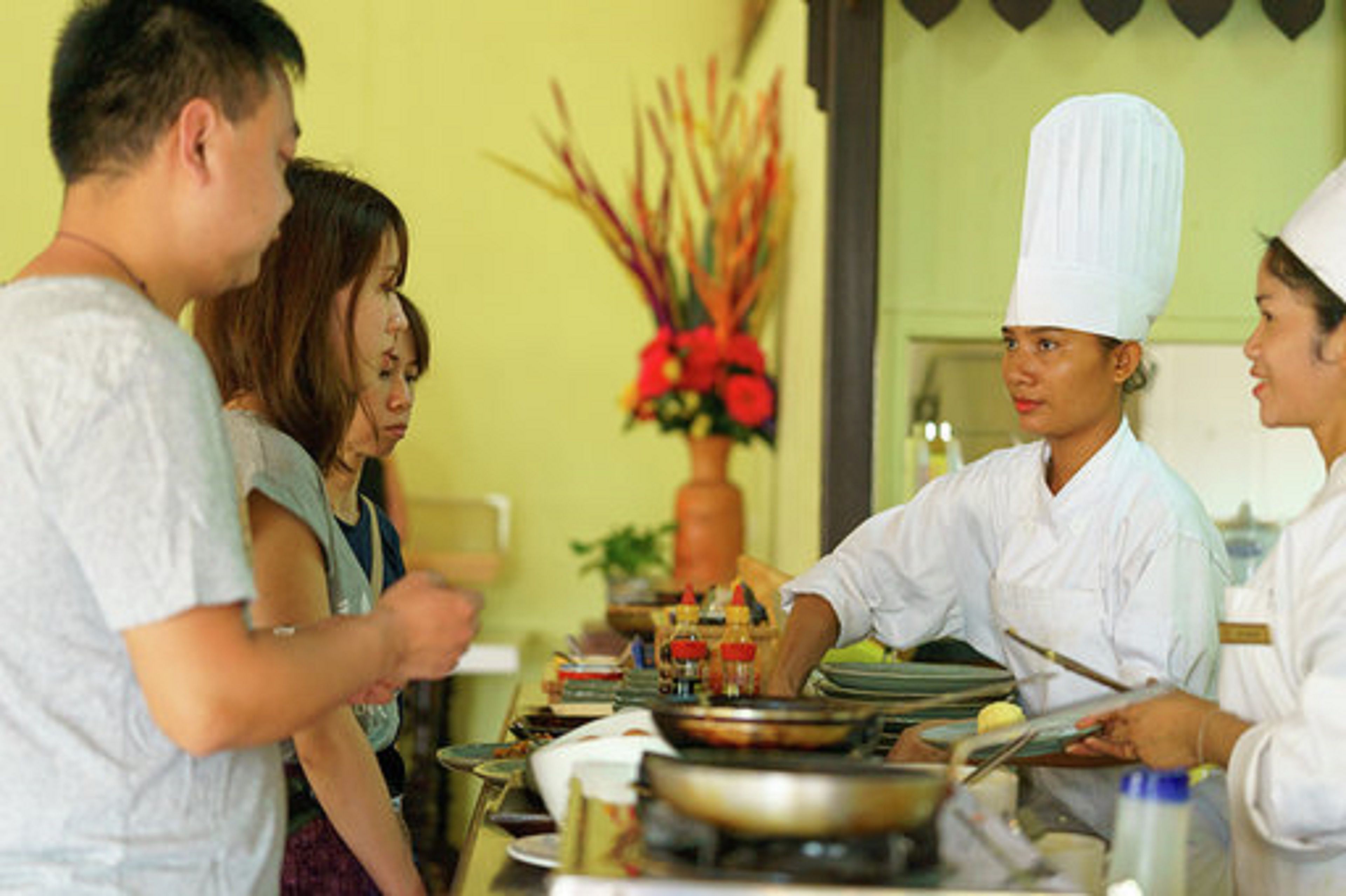 A group of people in a kitchen setting with a chef wearing a white hat, a dining table with various dishes, and a vase with red flowers in the background.