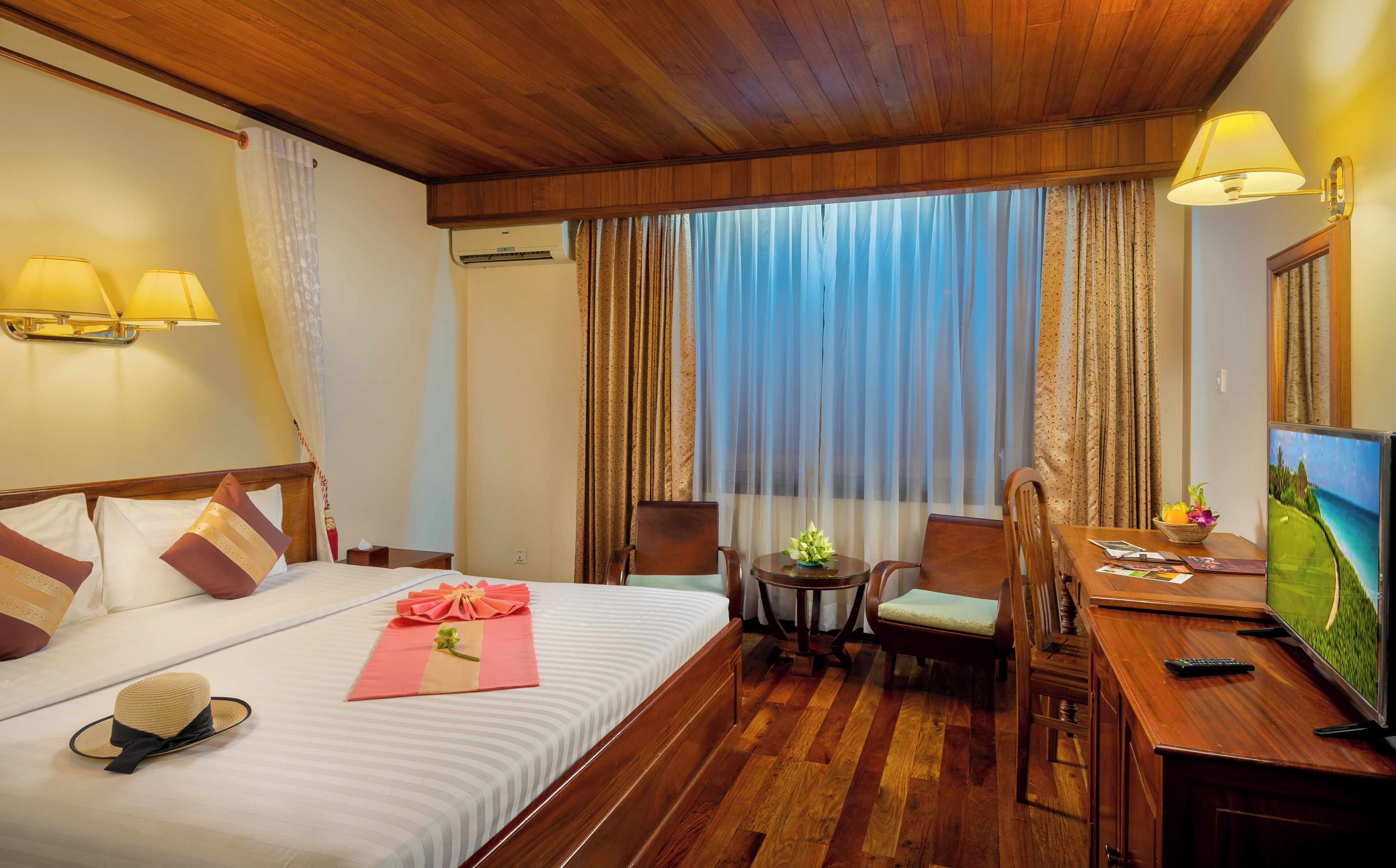 A hotel room with a bed, bedside lamps, a desk with a chair, a television, and a window with curtains.