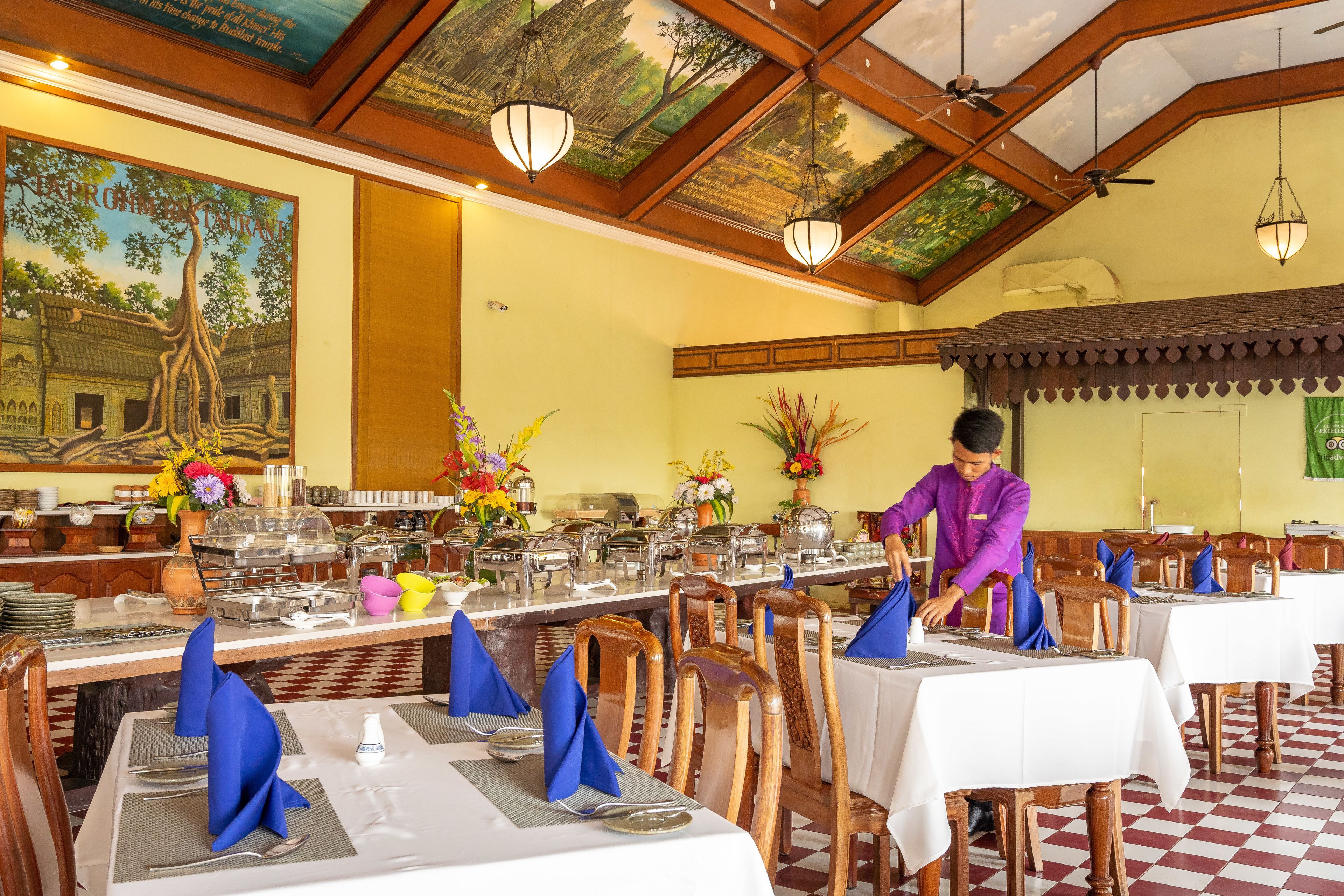 A restaurant with wooden tables, blue napkins, and a waiter in a purple uniform.