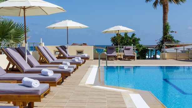 A poolside area with sun loungers and umbrellas, a clear blue sky, and a palm tree. - Panorama Hotel Apartments (Hersonissos)