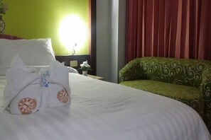 Een net opgemaakt bed met kussens en een opgevouwen deken, een armstoel met groen patroon, en een muur met een klok en een lamp.