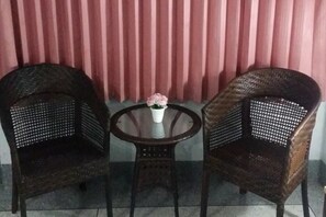Twee rieten stoelen en een kleine ronde tafel met een witte vaas en roze bloemen.