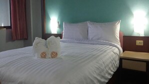 Ein ordentlich bezogenes Bett mit weißen Laken und zwei Kissen, ein Nachttisch aus Holz mit einer Lampe und eine wandfarbene Fläche in Türkis.