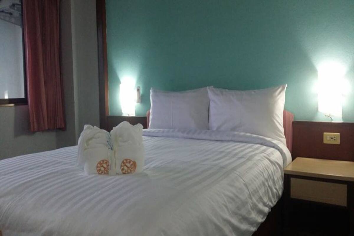 Un lit bien fait, avec du linge de lit blanc et deux oreillers, une table de chevet en bois avec une lampe, et un mur de couleur turquoise.
