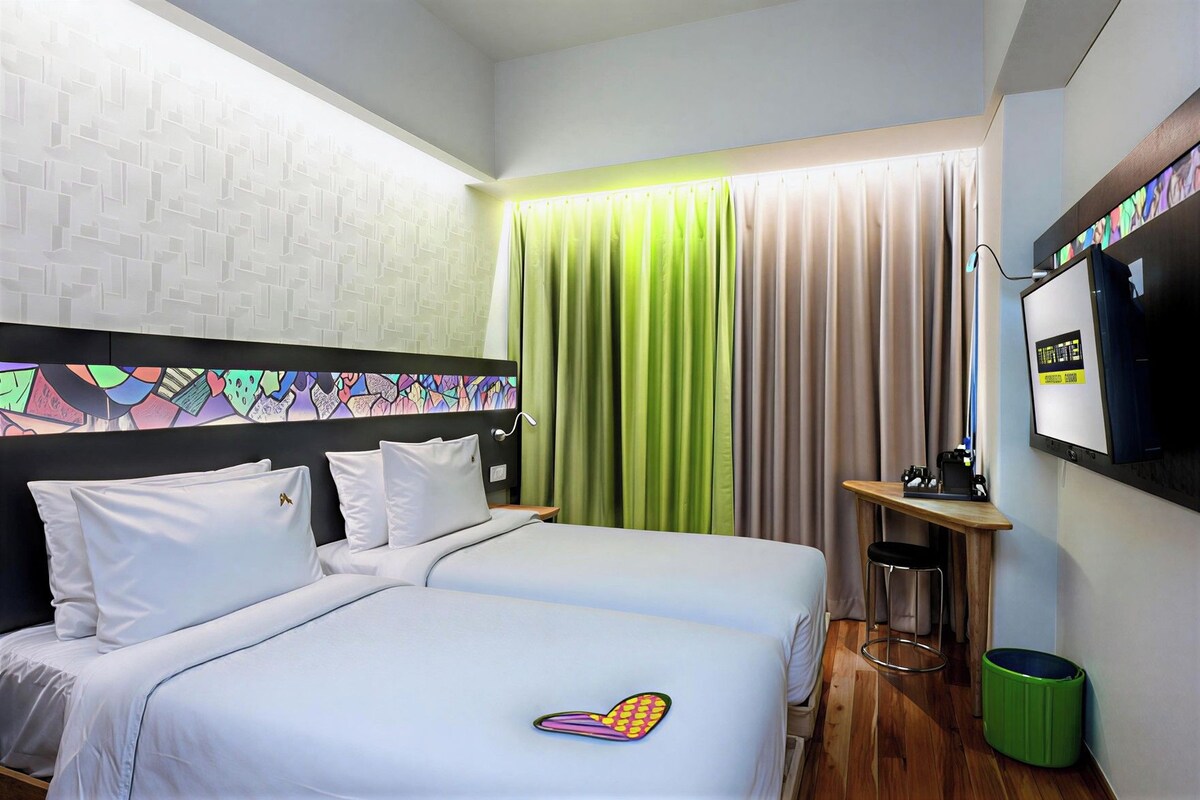 A hotel room with a bed, a desk, a television, and a green trash bin.