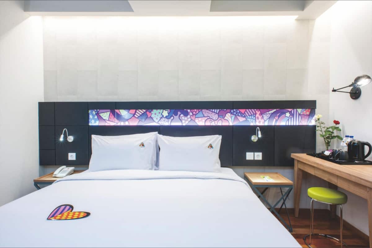 Ein Hotelzimmer mit einem Bett, einem Schreibtisch, einem Stuhl, einem Nachttisch, einer Lampe und einem Wandbild.