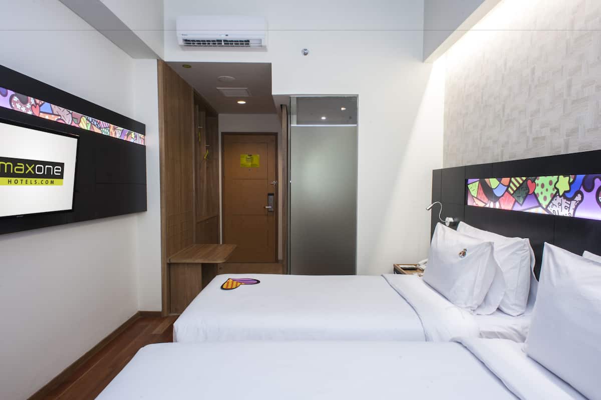 Ein Hotelzimmer mit zwei Betten, einer Holzbank, einer Tür und einem an der Wand befestigten Fernseher, der den Namen des Hotels zeigt.