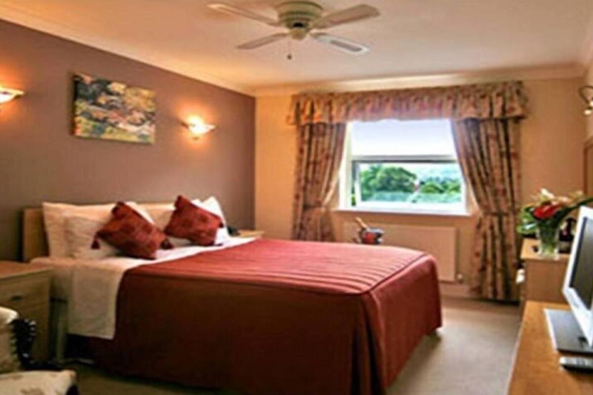 A hotel room with a bed, a desk with a computer, a window with curtains, and a ceiling fan.