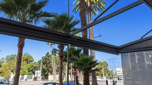 An outdoor seating area with tables and chairs, palm trees, and a view of a street with cars and pedestrians. - Malak Hotel (Rabat)