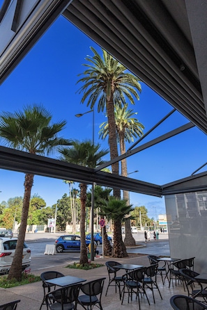 An outdoor seating area with tables and chairs, palm trees, and a view of a street with cars and pedestrians. - Malak Hotel (Rabat)