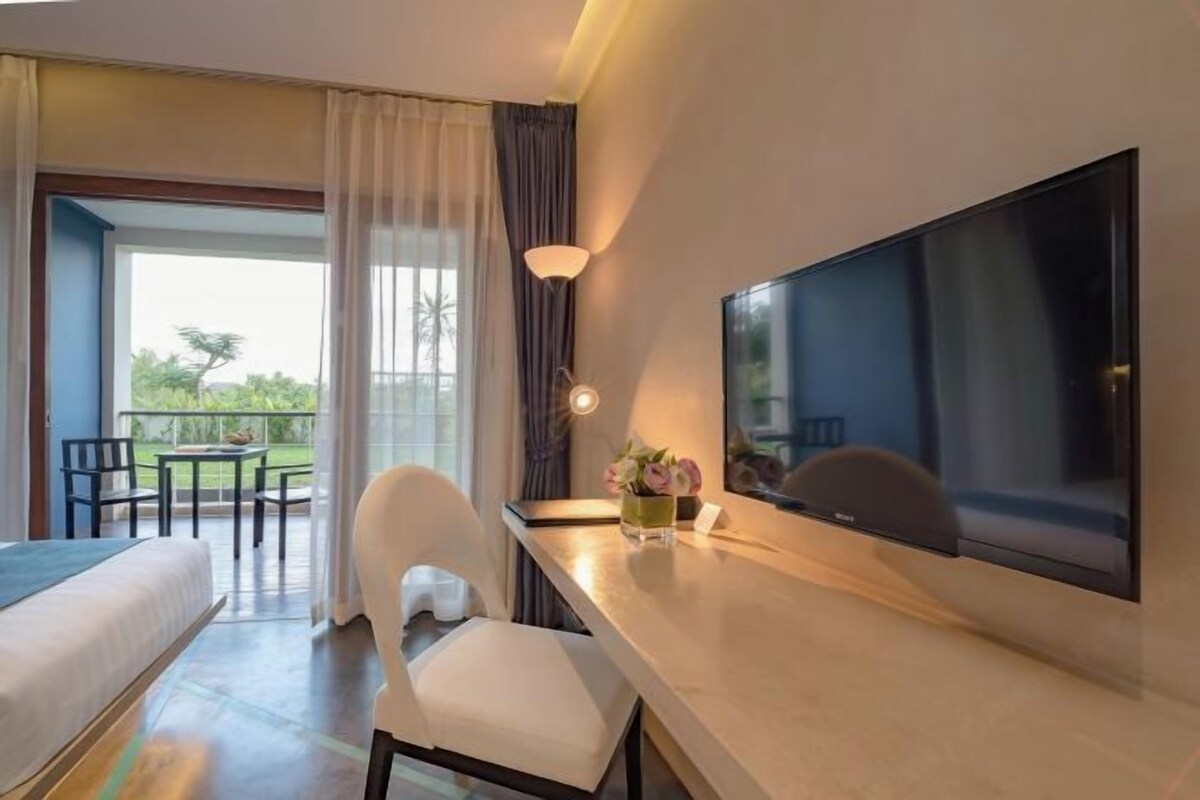 A hotel room with a bed, a desk with a flat-screen TV, a chair, a small table with a vase of flowers, and a view of the outdoors through a large window.