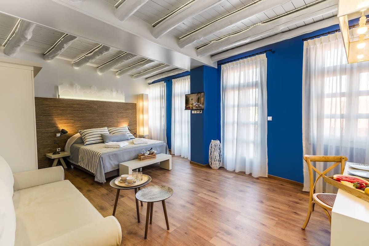 Un dormitorio moderno con cabecera de madera, una cama con ropa de cama a rayas, una mesita de noche, una pequeña mesa redonda con una bandeja y una silla con una cesta de frutas.
