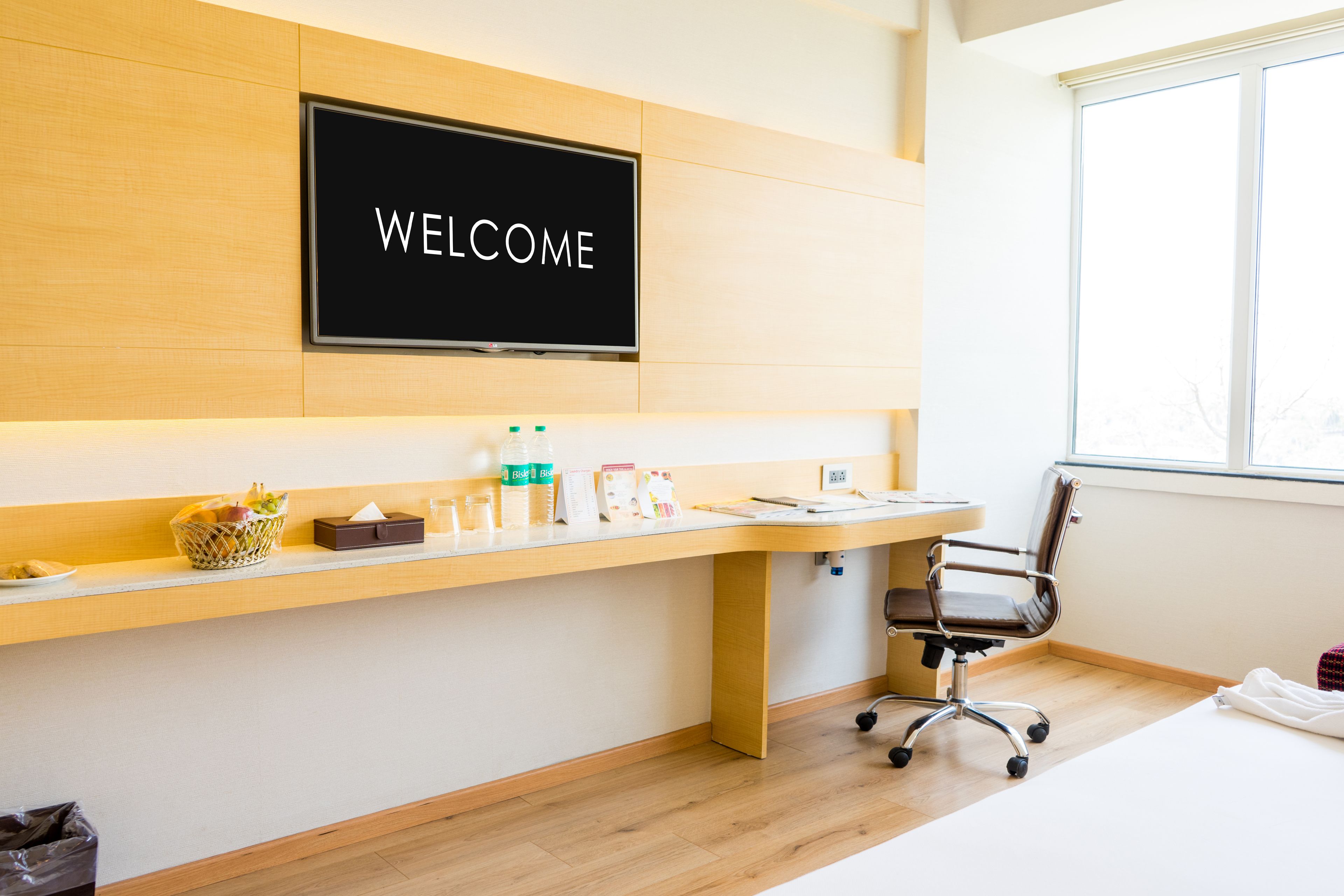 A modern hotel reception area with a "WELCOME" sign, a desk with various items, a chair, and a window providing natural light.