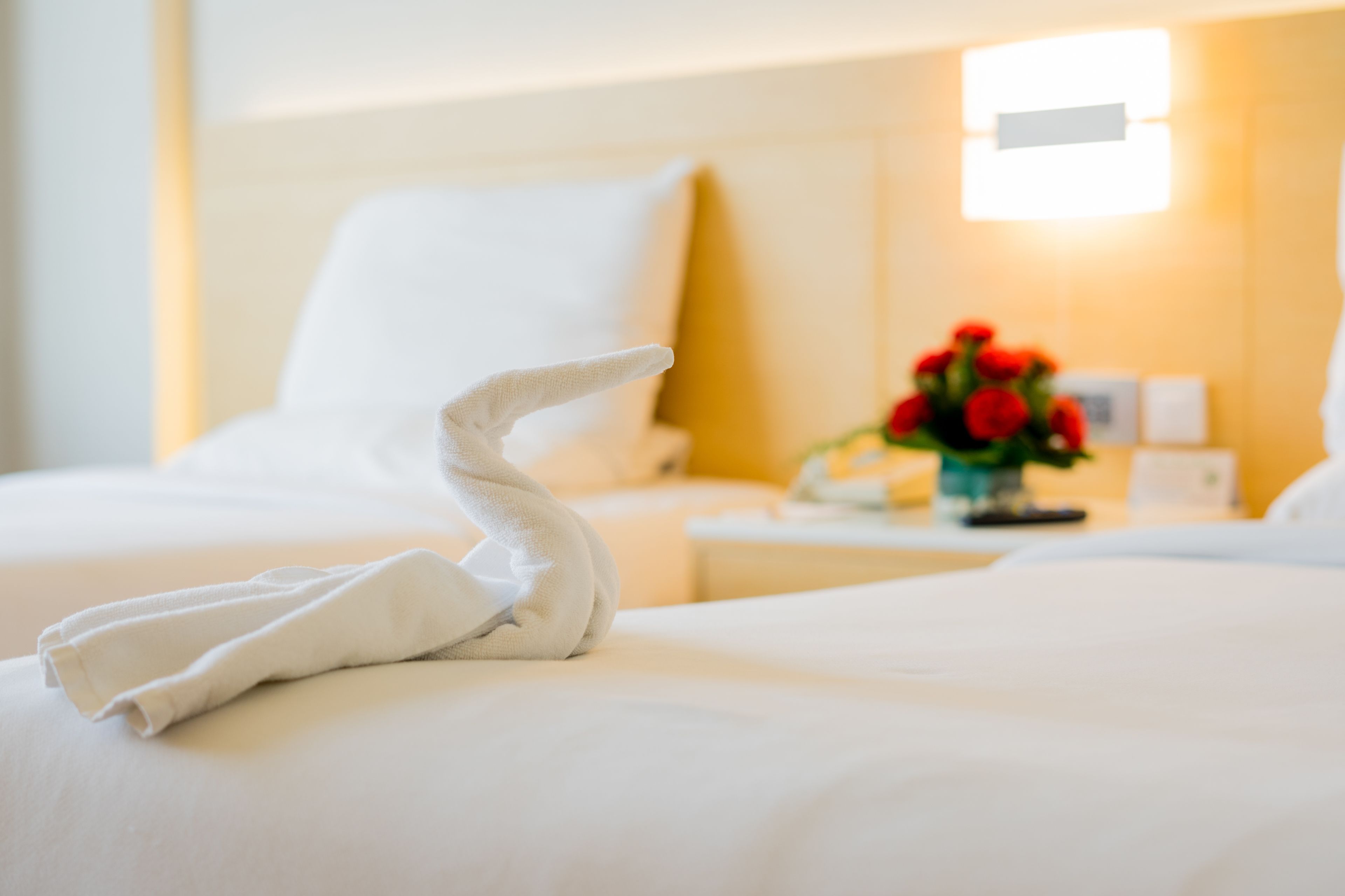 A neatly made bed with a towel folded into a swan shape, a white pillow, and a nightstand with a vase of red flowers.