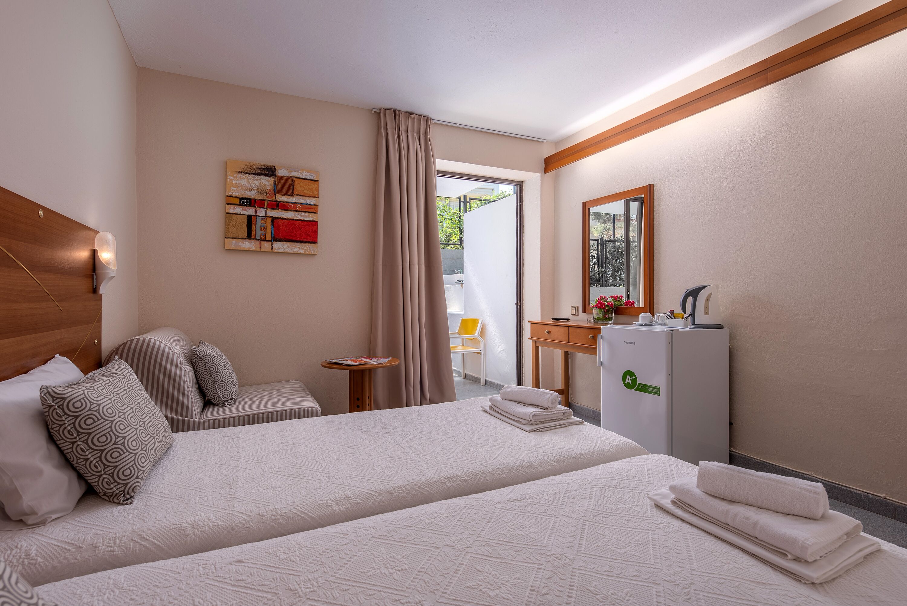 A hotel room with two beds, a small table, a desk with a mirror, and a refrigerator.