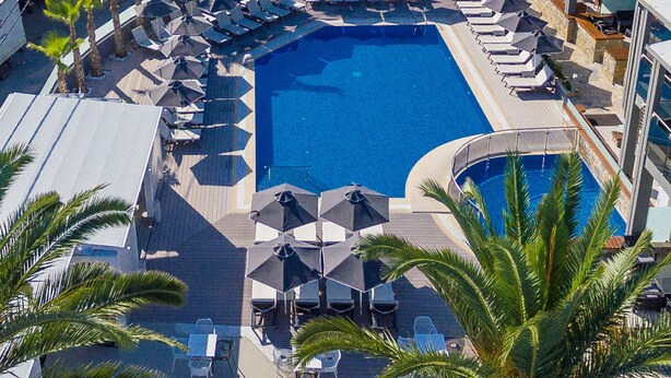 Vista aérea de un hotel con piscina central rodeada de tumbonas y sombrillas, palmeras y una estructura de edificio moderna al fondo.