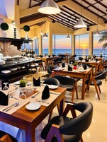 Un restaurante con mesas dispuestas para comer, una zona de bufé y vistas a la puesta de sol sobre el océano.