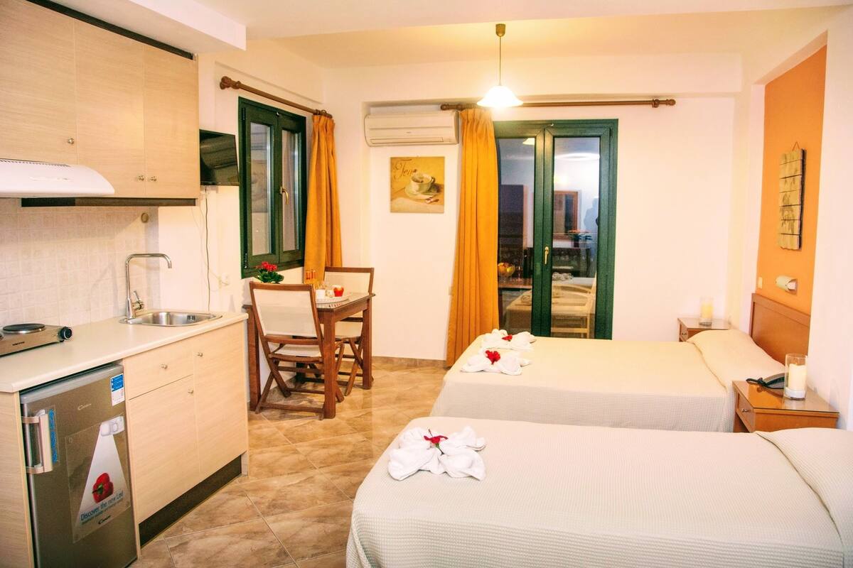 A hotel room with two beds, a kitchenette, a dining area, and a view of the cityscape.