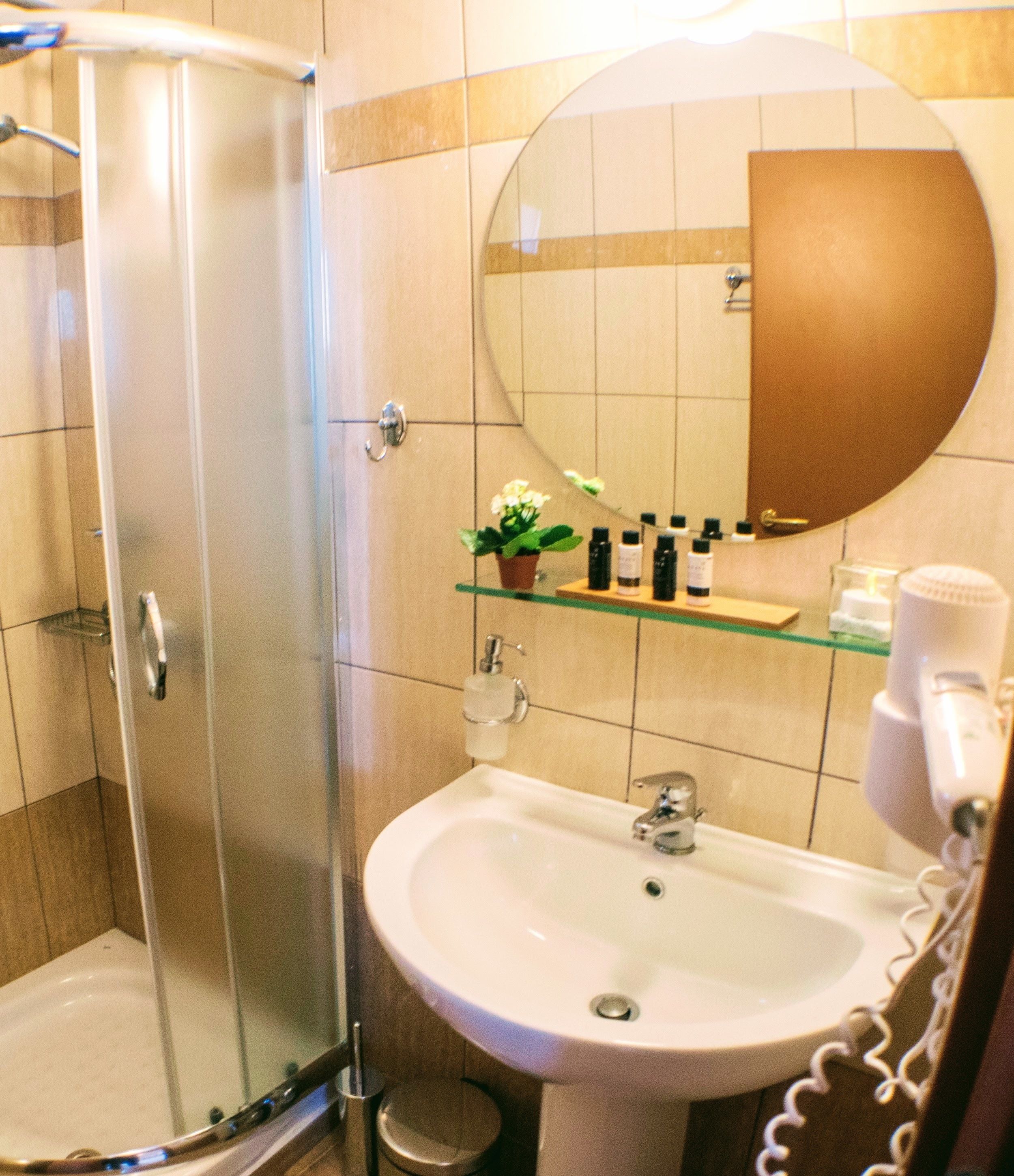 A bathroom with a glass shower enclosure, a white sink, a round mirror, and a shelf with toiletries.