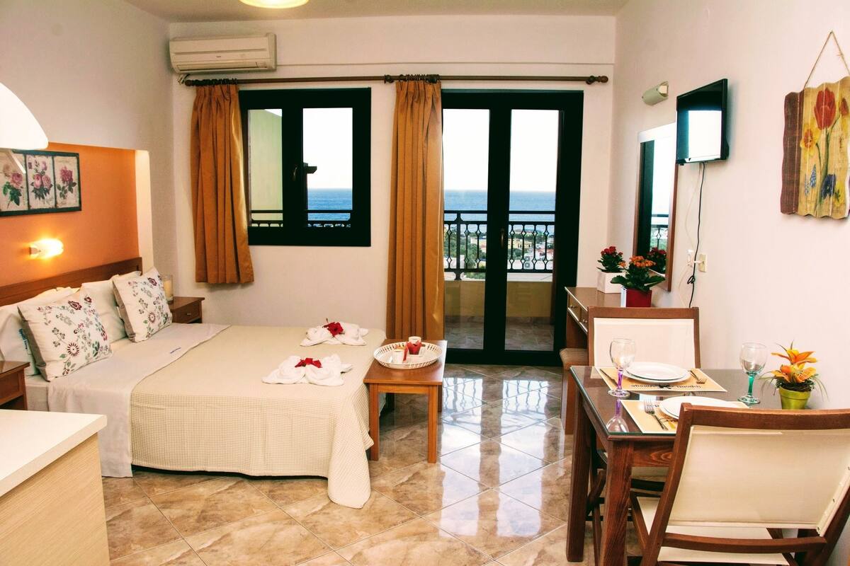 A hotel room with a bed, a dining table set for two, a small table with a vase of flowers, and a view of the sea through the windows.