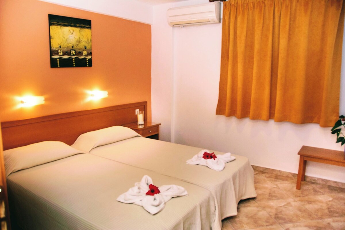 A hotel room with two beds, a nightstand, a wall-mounted air conditioner, and a window with curtains.