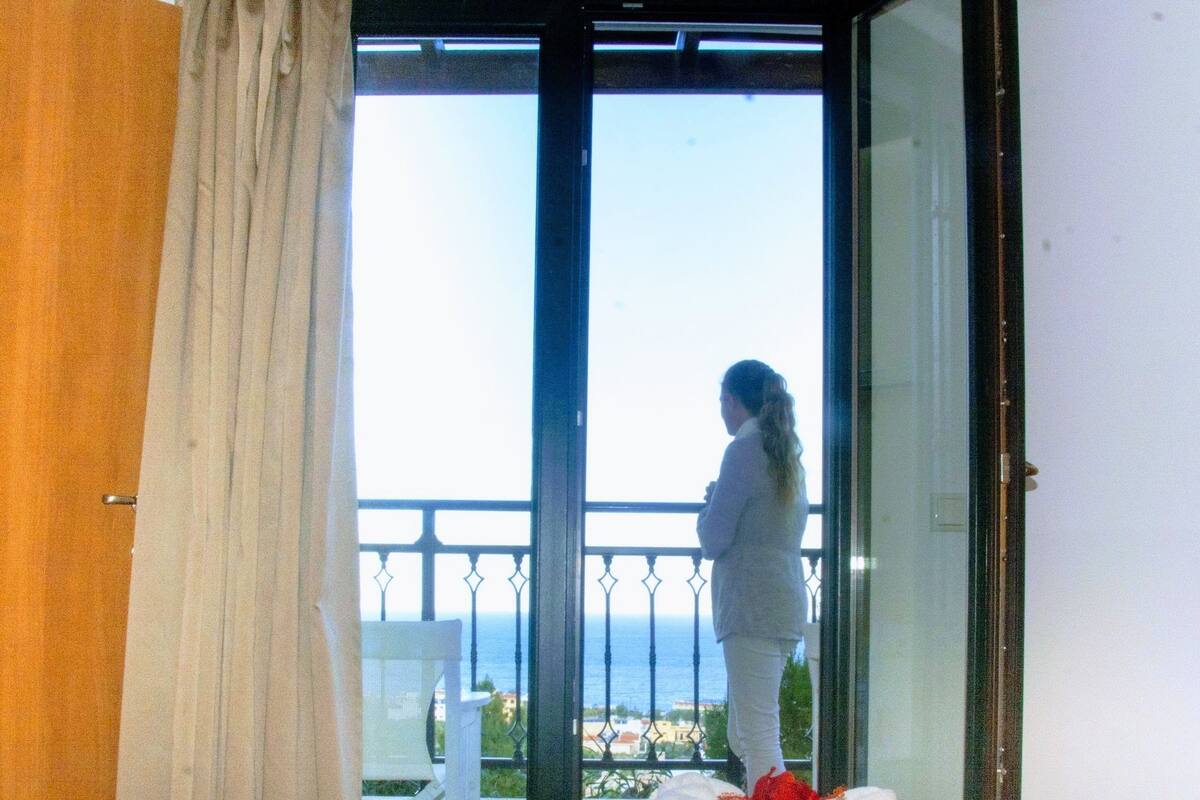 A room with a bed, a window with a view of the sea, and a curtain.
