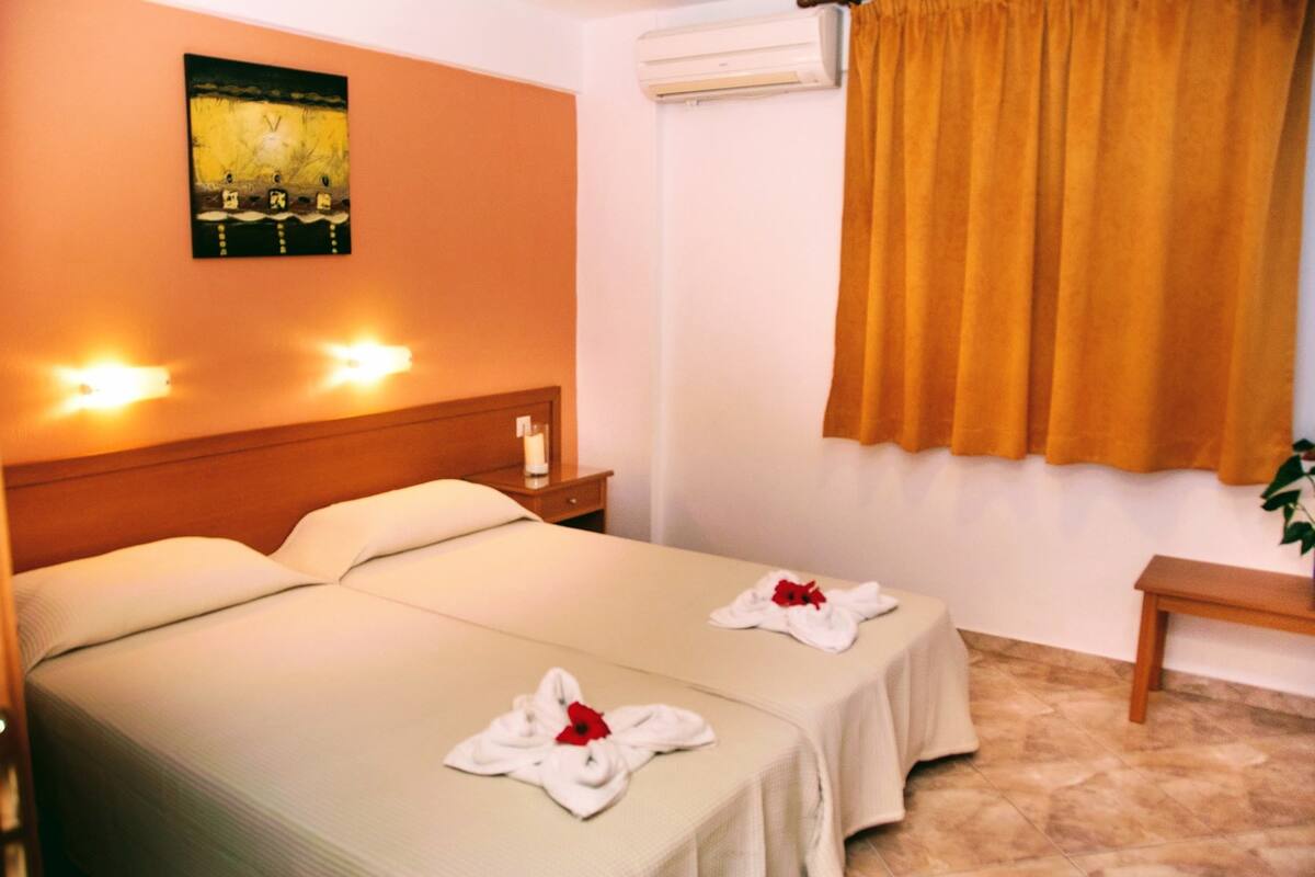 A hotel room with two beds, a wooden headboard, a wall-mounted air conditioner, a window with curtains, and a bedside table with a lamp.