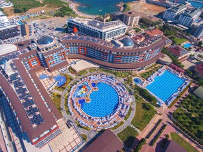 Oversigtsbillede af et resort med flere swimmingpools, inklusive en stor central pool, og forskellige bygninger herunder et hotel med en buet facade.