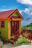 Um edifício pequeno e colorido com telhado vermelho e paredes verdes, apresentando uma porta vermelha e um pequeno alpendre com escadas de madeira.