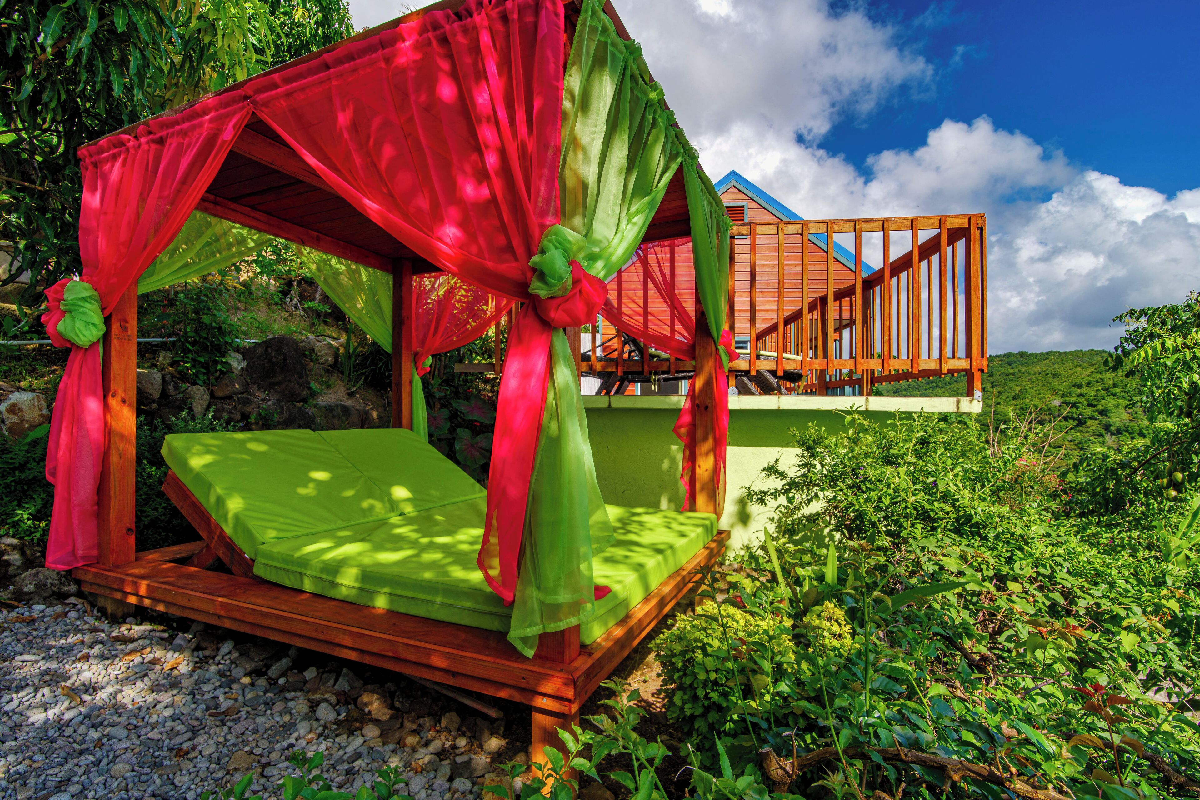 Una cama de madera con colchón verde y dosel rojo, frente a un fondo de vegetación exuberante y cielo azul despejado.