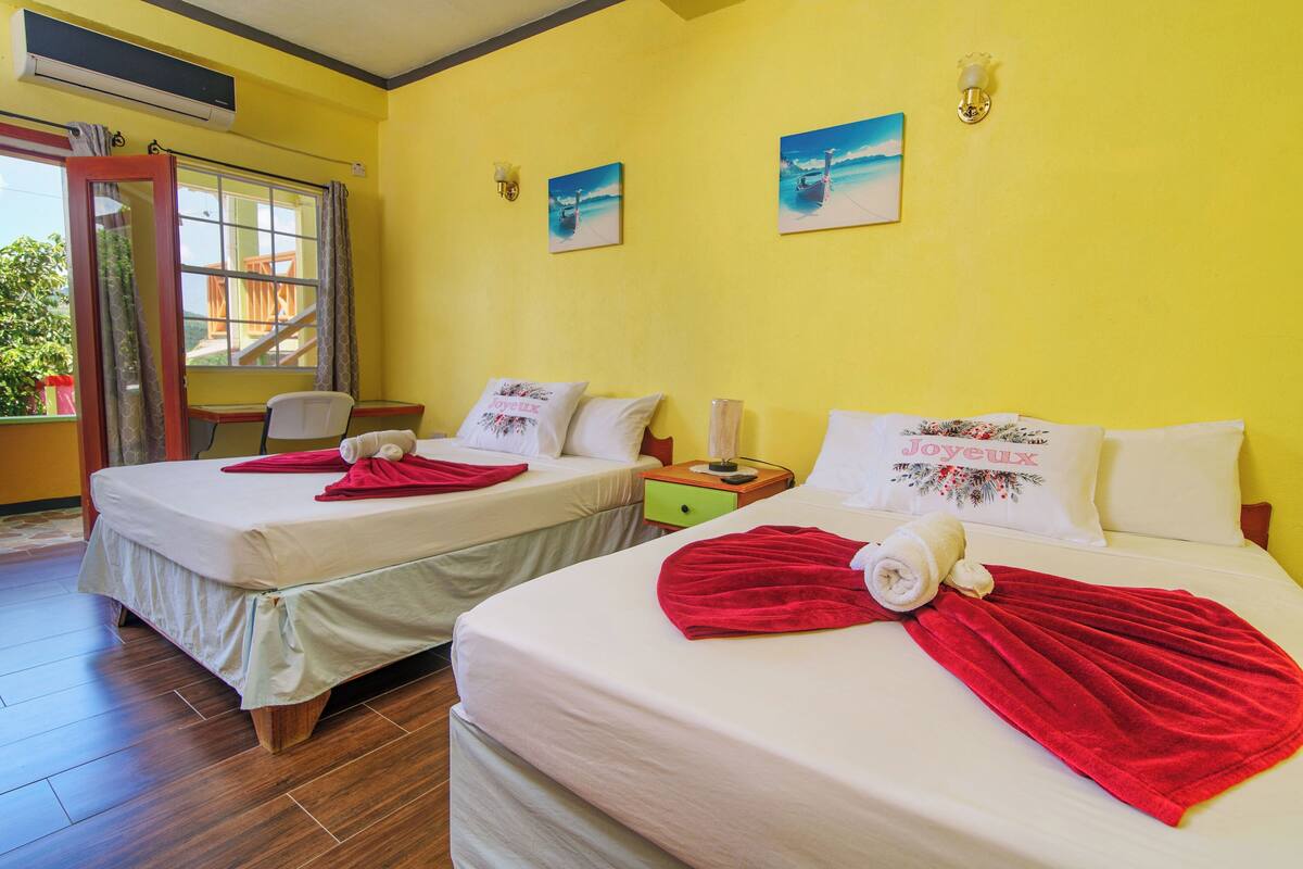 Two twin beds with white linens and red towels, a small wooden nightstand, a green drawer, and a window with curtains.