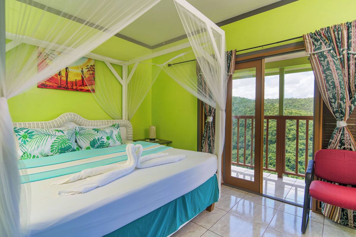 A bed with white linens and a canopy, a red chair, and a balcony with a view of greenery.