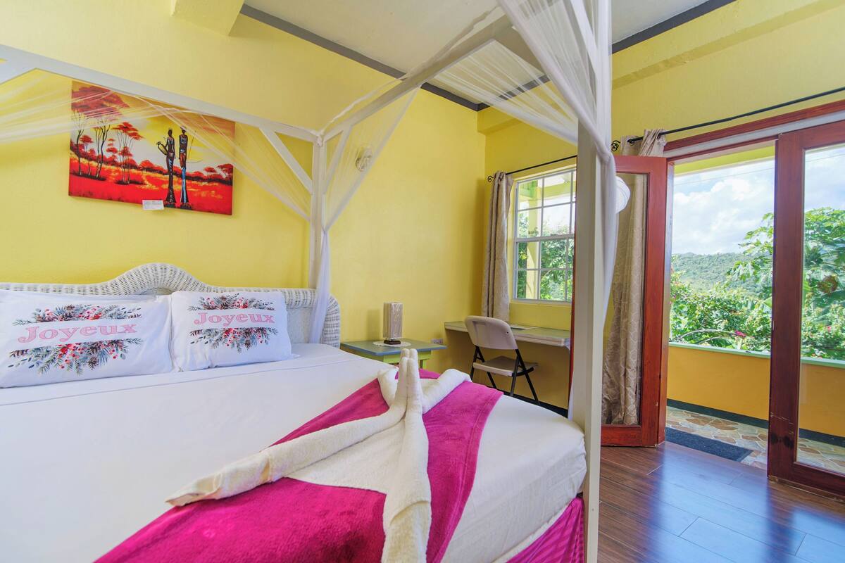 A bedroom with a bed, a desk, and a window with a view of greenery.