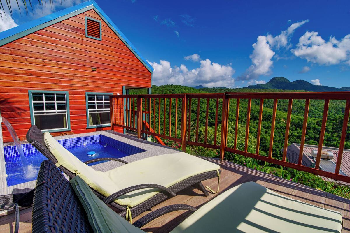 A wooden house with a hot tub and lounge chairs on a balcony overlooking a lush landscape.
