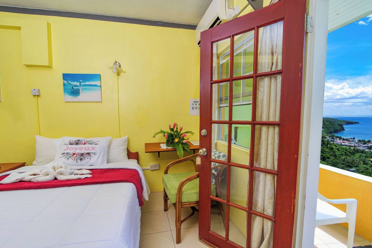A room with a bed, a chair, a small table with a plant, and a door leading to a balcony with a view of the sea.