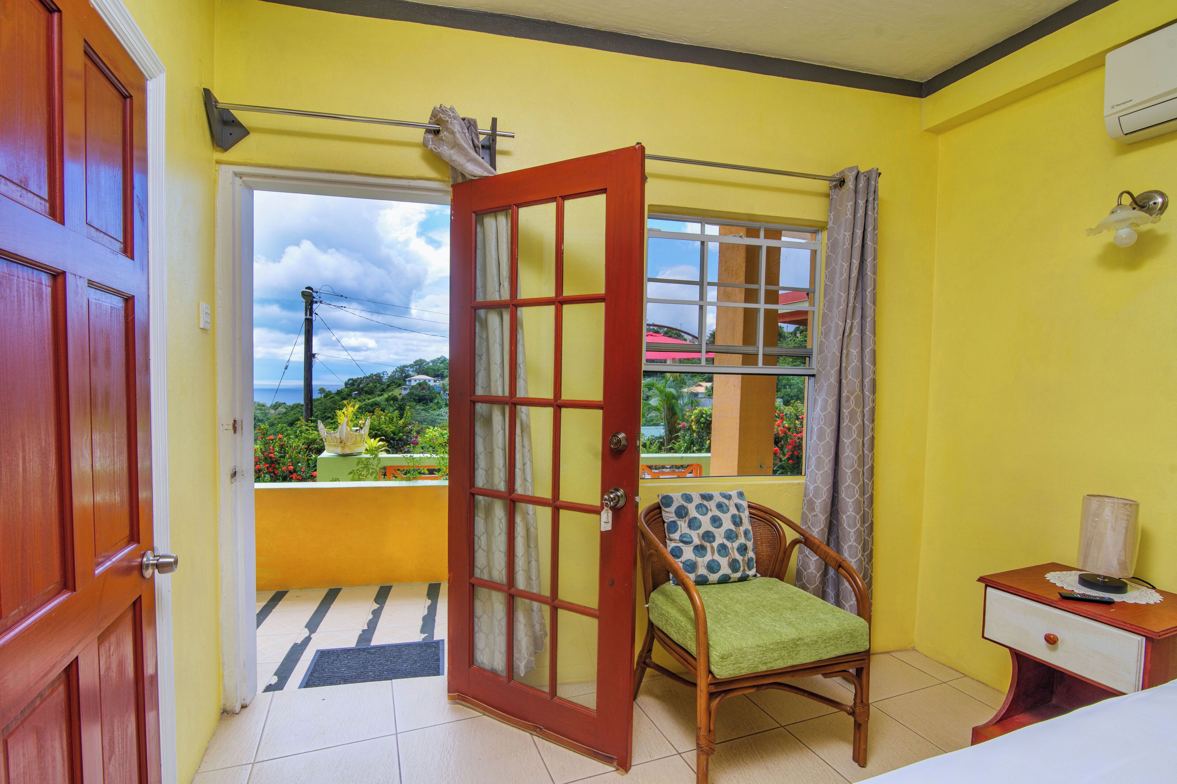 A room with a yellow wall, a wooden door, a window with a view of greenery, a chair with a cushion, and a bedside table with a lamp.