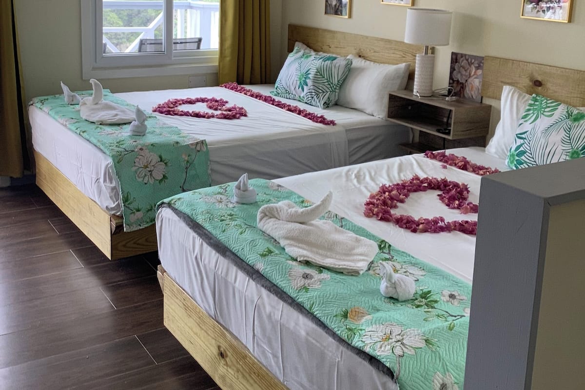 A hotel room with two beds, a window with curtains, a nightstand, a lamp, and framed floral artwork on the wall.
