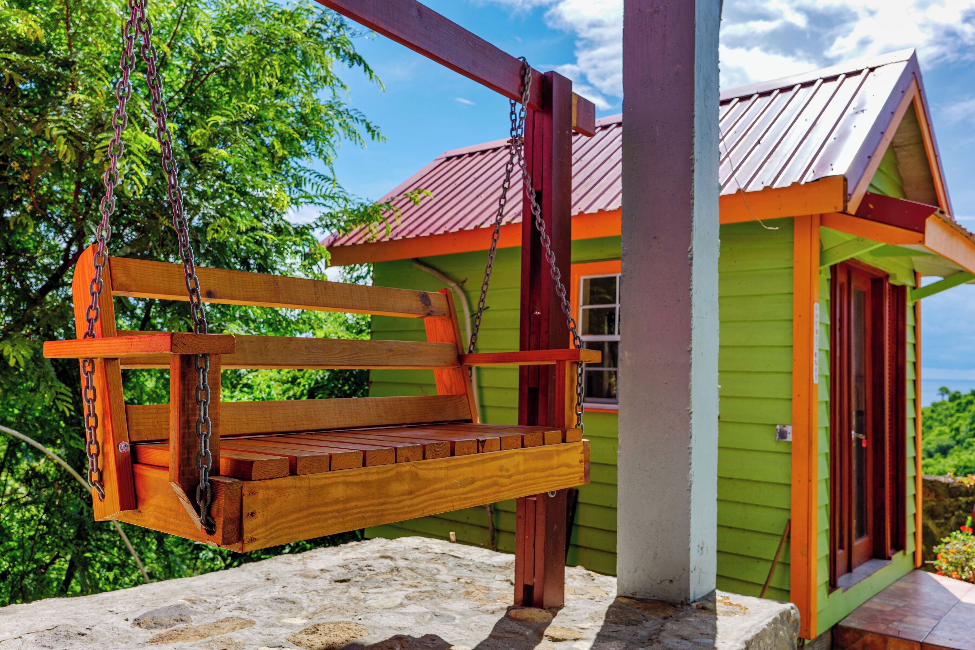 A wooden swing suspended from a metal frame, attached to a building with a green exterior and a red roof.