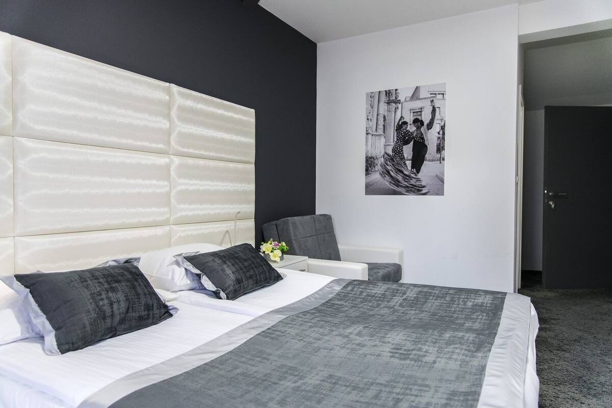 A modern hotel room with a large bed, a grey upholstered headboard, a bedside table with a vase of flowers, and a framed black and white photograph on the wall.