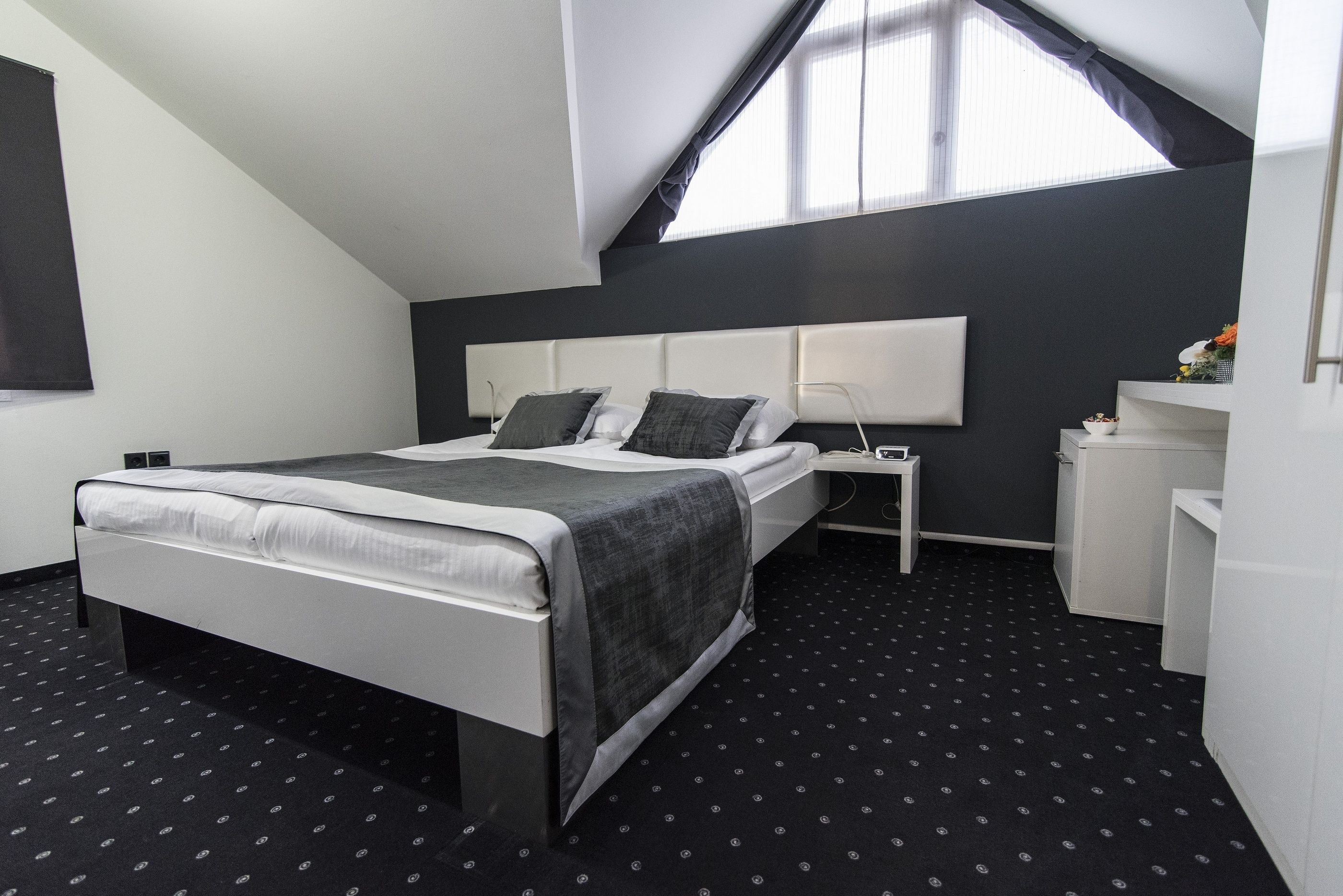 A modern hotel room with a bed, bedside table, and a small desk with a lamp.