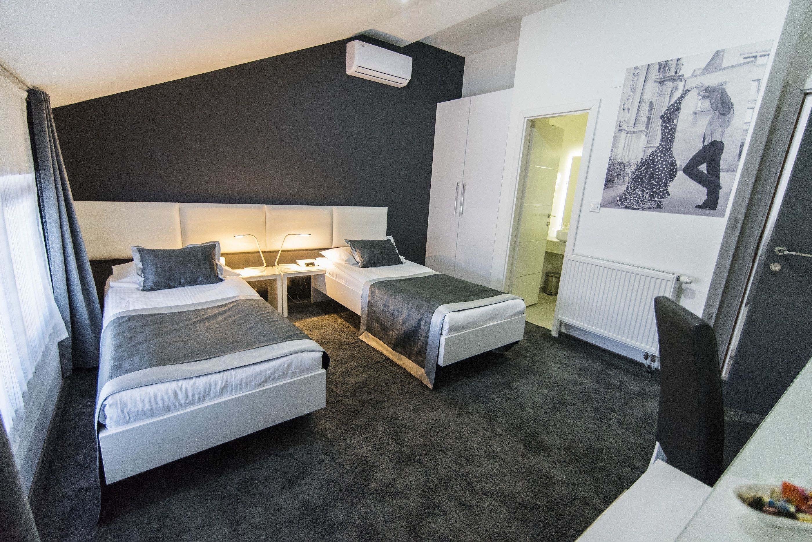 A modern hotel room with two single beds, a desk, a chair, a radiator, and a wall-mounted air conditioner.