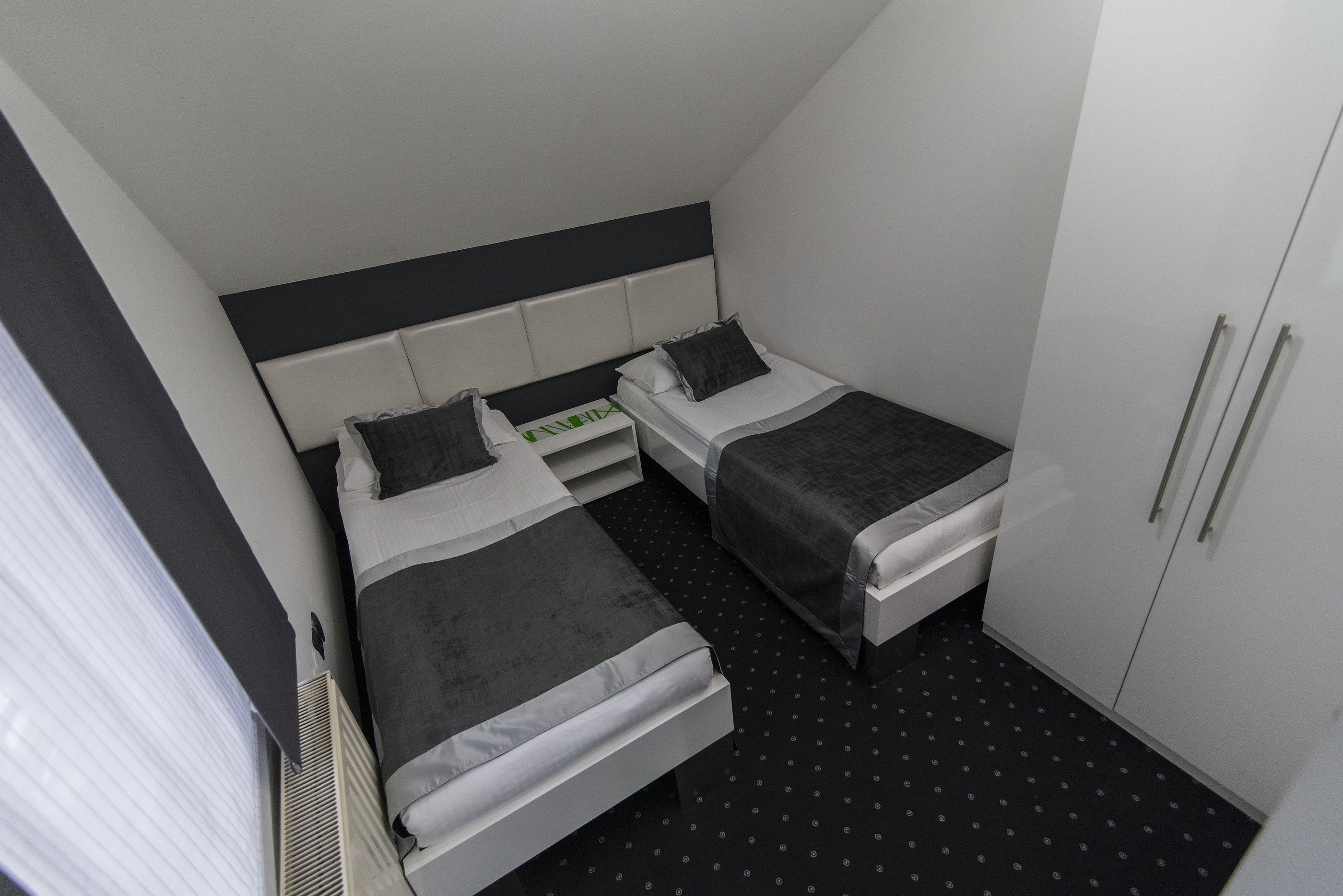 A small, modern hotel room with two single beds, a white wardrobe, and a black patterned floor.