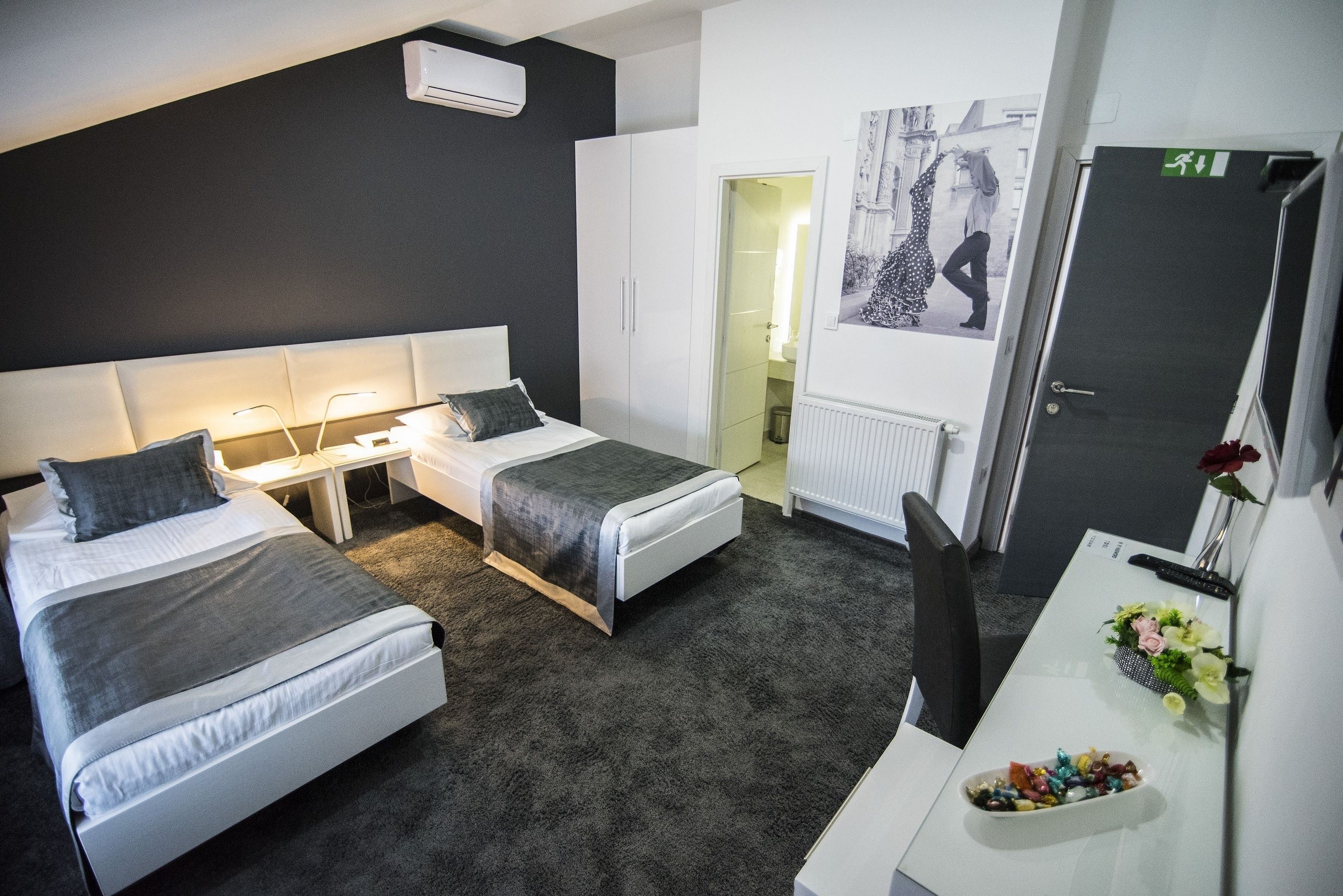 Una habitación de hotel moderna con dos camas individuales, un escritorio con computadora, una silla y una pequeña mesa con flores.