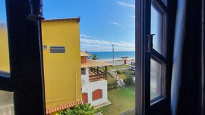 A view from a window overlooking a building with a yellow exterior and a white facade, a small patio area, and a beach in the background. - Pousada Pratagy (Saquarema)