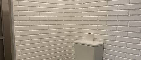 A bathroom with a white brick wall, a white toilet, a blue trash bin, and a shower area.