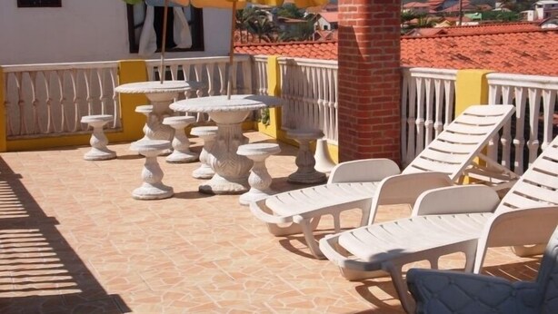 A terrace with white lounge chairs and tables, a brick pillar, and a view of a town with red-roofed houses. - Pousada Pratagy (Saquarema)