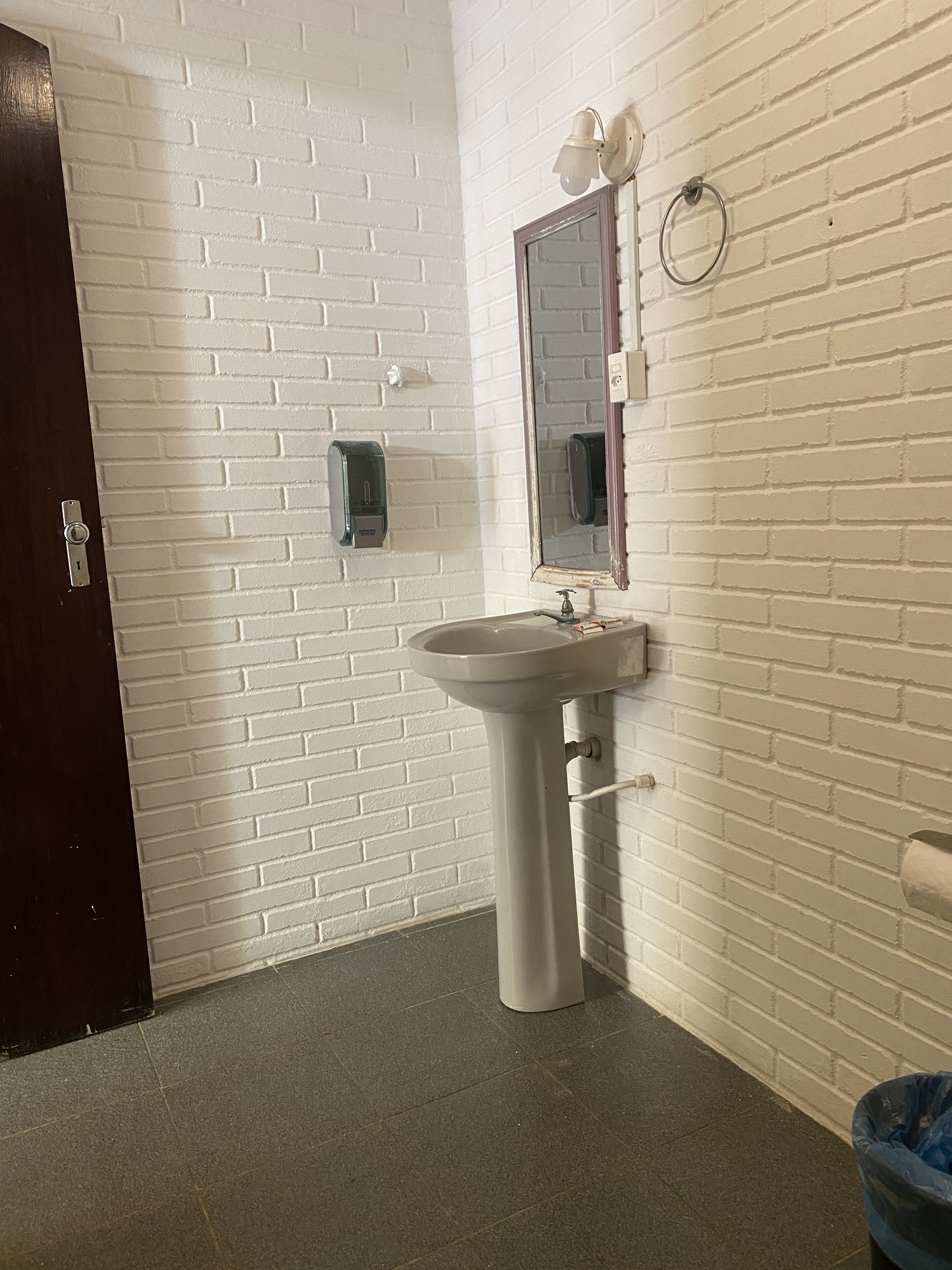 Un baño con pared de ladrillo blanco, un lavamanos, un espejo, un dispensador de jabón y un cesto de basura.