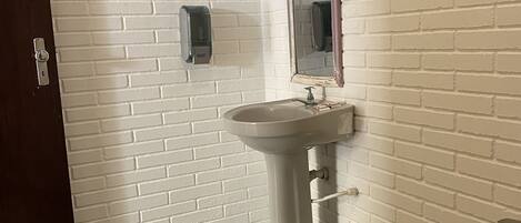 Un baño con pared de ladrillo blanco, un lavamanos, un espejo, un dispensador de jabón y un cesto de basura.