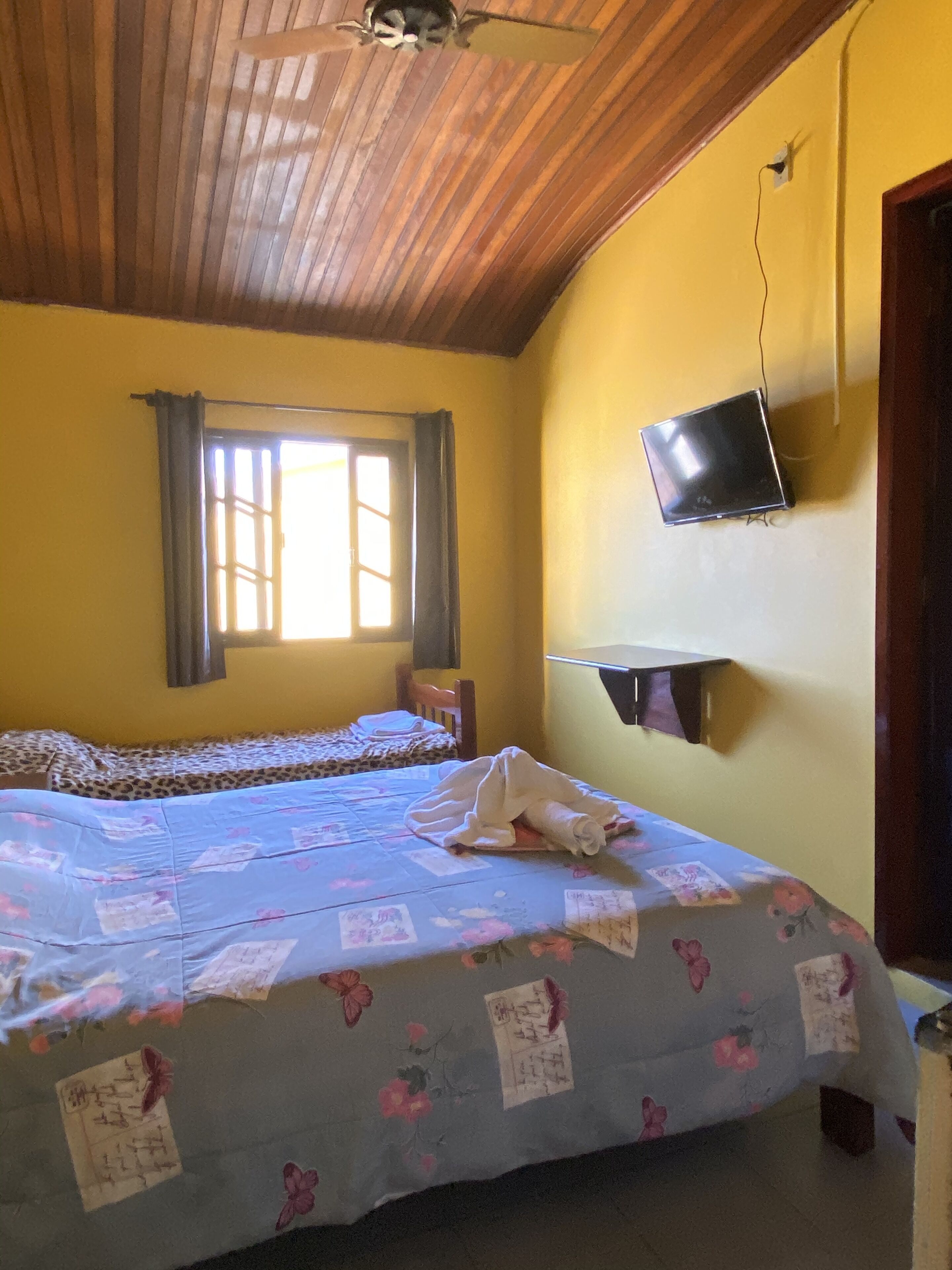 Una camera da letto con due letti, una televisione fissata al muro e una finestra con tende.