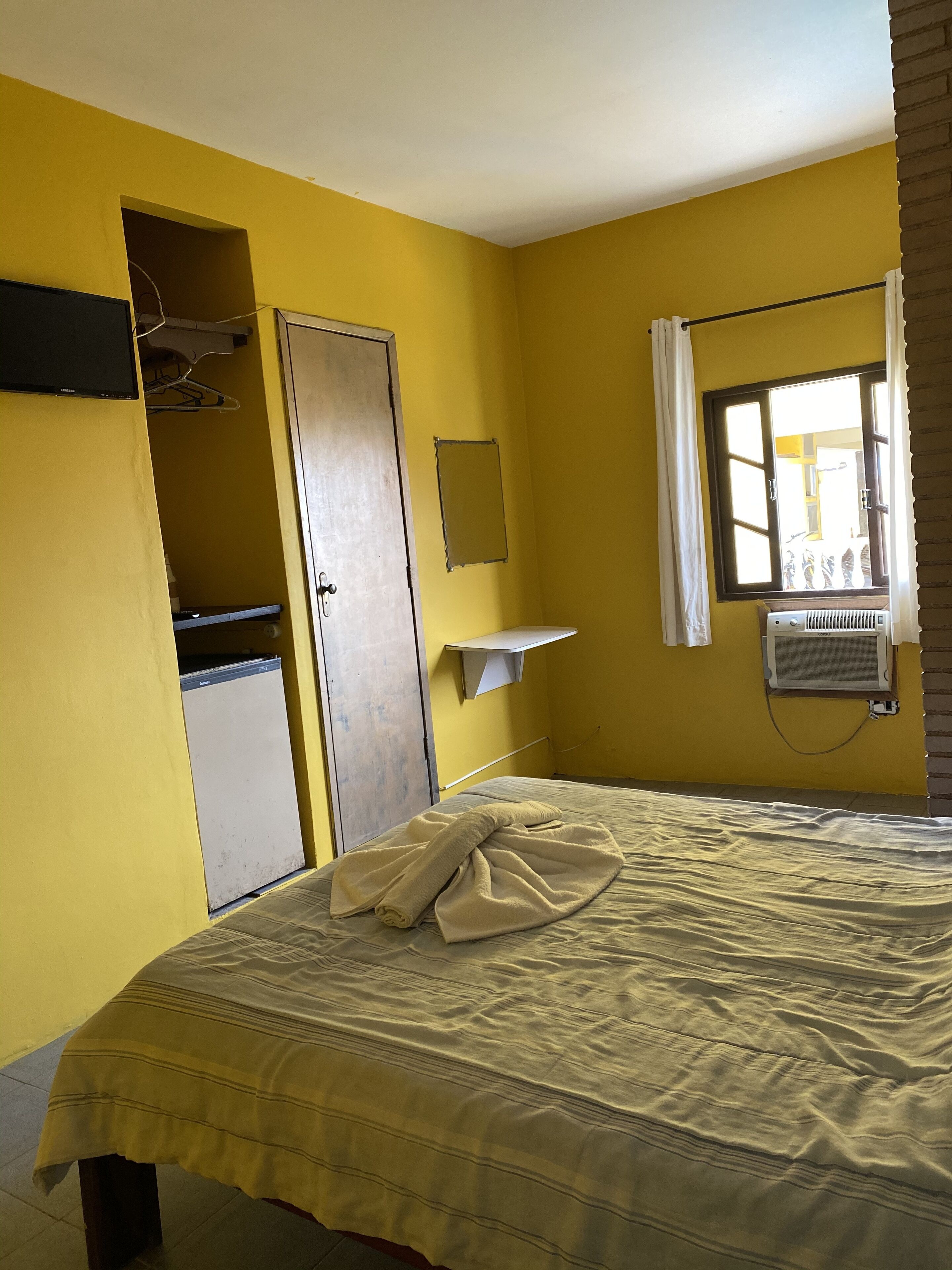 Una habitación con paredes amarillas, una cama con manta color beige, un armario, un estante pequeño y una ventana con cortina.